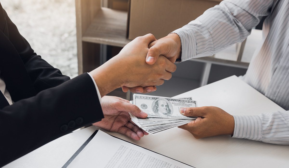 Two corporate businessmen shaking hands while one man places money on document in office room with corruption concept.